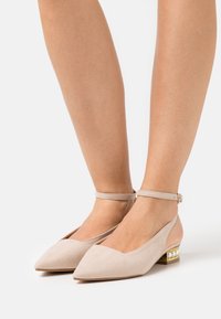 Mujer vistiendo unas bailarinas de punta beige con correas al tobillo y tacones bajos cuadrados decorados con perlas sobre un fondo blanco.