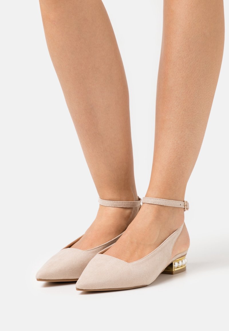 Mujer vistiendo unas bailarinas de punta beige con correas al tobillo y tacones bajos cuadrados decorados con perlas sobre un fondo blanco.