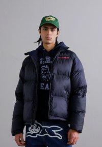 Schwarze Pufferjacke mit Reißverschluss und Seitentaschen, getragen über einem dunkelblauen Hoodie mit weißem Text, kombiniert mit blauen Jeans mit Grafik.