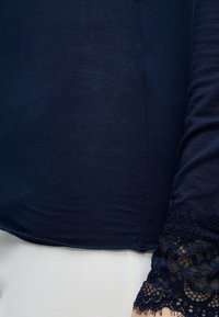 Μπλε ναυτικό τοπ με μακριά μανίκια και δαντελωτά μανσέτα, σε συνδυασμό με λευκό παντελόνι, δείχνοντας το κάτω μέρος του κορμού και το χέρι.