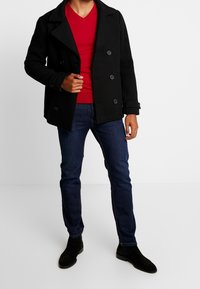Czarny płaszcz peacoat z podwójnym zapięciem na guziki, czerwony sweter v-neck, ciemnoniebieskie dżinsy i czarne botki za kostkę, tworzące warstwowy strój.