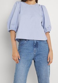 Kvinna iklädd en ljusblå blus med puffärmar och högmidjade blå jeans, stående mot en enkel ljus bakgrund.