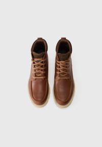 Un par de botas de cuero marrón con cordones, cuellos acolchados y suelas de goma beige claro sobre un fondo blanco.