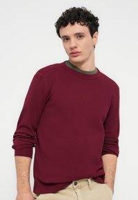 Μακρυμάνικη μπλούζα μπουρντό με ριμπ υφή, στρογγυλή λαιμόκοψη και διακριτική λεπτομέρεια λογότυπου. Φορεμένη πάνω από σκούρο πράσινο πουκάμισο, σε συνδυασμό με χακί παντελόνι.