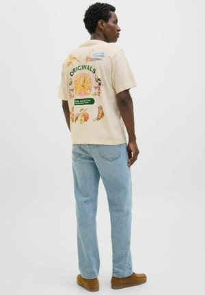 Beige Baumwoll-T-Shirt mit bunten Grafiken auf dem Rücken, darunter Blumen und ein Oktopus, kombiniert mit hellblauen Jeans.