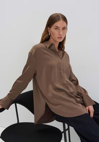 Bruine satijnen blouse met lange mouwen, knoopsluiting bij de kraag en een gebogen zoom, gecombineerd met donkere spijkerbroeken, zittend op een zwarte stoel.