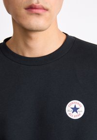 Sudadera de algodón negra con un parche de logo redondo que presenta un diseño de estrellas en rojo, blanco y azul, cuello cosido y textura simple, sin adornos.