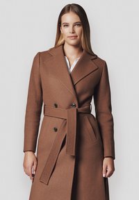 Brązowy płaszcz z mieszanki wełny z paskiem w talii, podwójnym zapięciem z przodu, szerokimi klapami oraz bocznymi kieszeniami. Gładka tekstura o subtelnej ciepłocie.