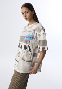 Przesadny, krótki T-shirt z krótkim rękawem w odcieniu beżowym z kratkowanym wzorem. Zawiera graficzne nadruki, w tym słowo "Zrelaksowany" w niebieskich i czarnych akcentach.