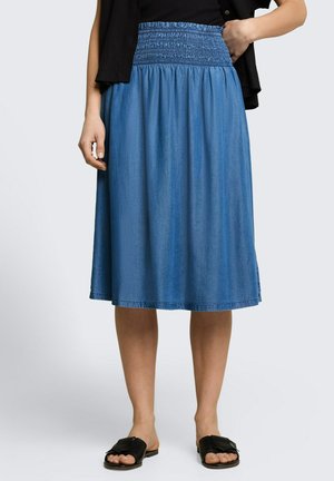 Vrouw draagt een knielange blauwe spijkerrok met een geplooide tailleband en zwarte slip-on sandalen, staand tegen een effen achtergrond.