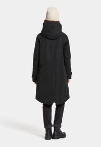 Lang zwart waterdicht jack met een capuchon, voorzien van een ronde zoom en verstelbare mouwen. Draagt een zwarte broek en een beige gebreide muts.