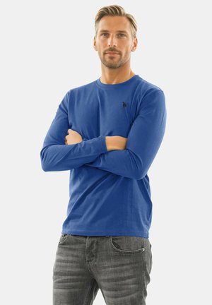 Man with folded arms wearing a blue long-sleeve shirt and gray jeans, standing against a plain light background.