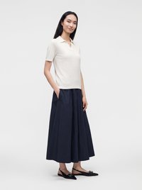 Femme aux longs cheveux foncés portant un polo blanc à manches courtes, une jupe midi plissée bleu marine et des ballerines à bride arrière noires, debout les mains dans les poches.