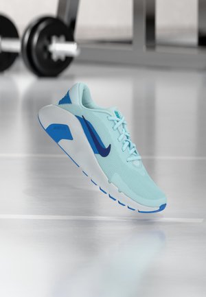 Pantof de alergare Nike albastru deschis cu swoosh și accente pe talpă de culoare albastru închis, prezentat în aer pe podeaua reflectorizantă a sălii de sport, cu echipamente de exerciții în fundal.
