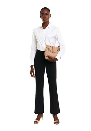 Vrouw in een witte blouse met knopen, zwarte broek en beige open teen hakken, die een beige handtas met gouden ketting vasthoudt tegen een witte achtergrond.