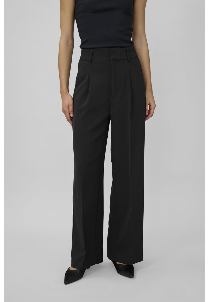 Mustia korkeavyötäröisiä housuja suoran lahkeen istuvuudella, joissa on poimuja ja sileä pinta. Yhdistettynä teräväkärkisiin mustiin kenkiin.