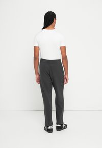 Muškarac s pletenicom, odjeven u bijelu majicu, tamnosive trenirke i crne tenisice, stoji okrenut prema bijelom zidu.