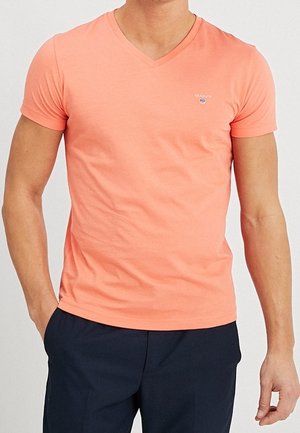 Mann trägt ein eng anliegendes korallenfarbenes T-Shirt mit V-Ausschnitt und kurzen Ärmeln sowie dunkle marineblaue Hose, steht mit einer Hand in der Tasche.