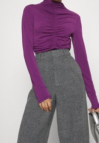 fialový top s dlouhým rukávem a nařasenými detaily v přední části, zkombinovaný s vysokým pasem širokými kalhotami s houndstooth vzorem. Ruka drží stříbrnou kabelku.