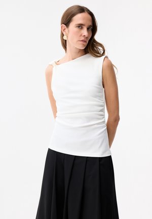 Vrouw met lang bruin haar, gekleed in een mouwloos wit gerimpeld topje met een asymmetrische halslijn en een zwarte geplooide rok, tegen een witte achtergrond.