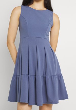 Sukienka bez rękawów do kolan w stonowanym niebieskim kolorze, z dopasowanym górnym partem, plisowanym pasem i rozkloszowanym, warstwowym dołem, noszona przez stojącą osobę.