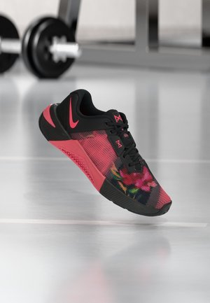 Scarpa sportiva nera e rossa con motivo floreale e logo Nike, esposta in un ambiente da palestra con manubrio sfocato sullo sfondo.