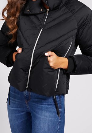 Kobieta ubrana w czarną pikowaną kurtkę z ukośnymi białymi zamkami i niebieskie dżinsy, z rękami w przednich kieszeniach.