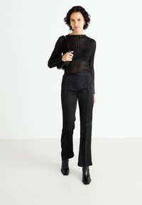 Schwarze transparente langärmelige Bluse mit vertikalen Streifen, kombiniert mit hochtaillierten schwarzen Satinhosen und spitzen schwarzen Ankle Boots.