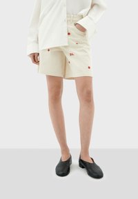Crème-kleurige shorts met rode geborduurde bloemen en de tekst "SOL", gecombineerd met een wit oversized shirt en zwarte instap schoenen.