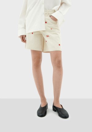 Krémové šortky s červenými vyšitými kvetinami a textom "SOL", spárované s bielou oversized košeľou a čiernymi slip-on topánkami.