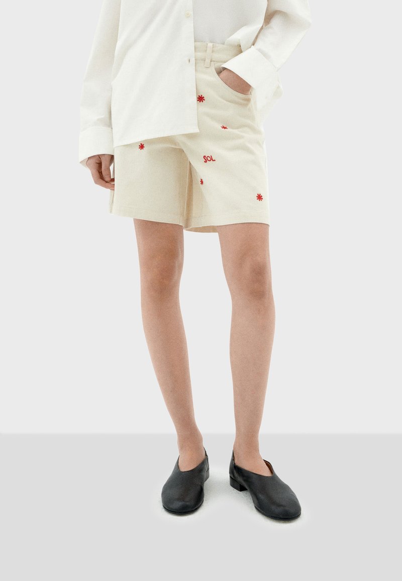 Crème-kleurige shorts met rode geborduurde bloemen en de tekst "SOL", gecombineerd met een wit oversized shirt en zwarte instap schoenen.