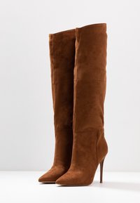 Stivali sopra il ginocchio in camoscio marrone con punta appuntita e tacco a stiletto. Caratterizzati da design senza cuciture e texture liscia.