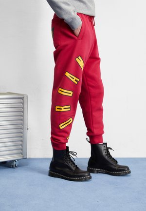 Person trägt rote Sweatpants mit gelbem "JORDAN" Schriftzug am Bein und schwarzen Schnürstiefeln, steht auf blauem Teppich neben einer Metallkiste.