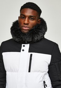 Padded jacket featuring a black and white color block design. Soft fur collar, front zipper with button closure, and zippered side pockets.