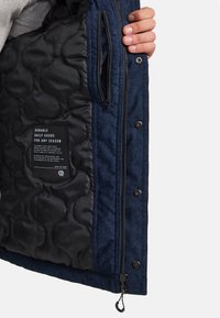 Blaue Cordjacke mit einer gesteppten schwarzen Fütterung, ausgestattet mit einem Frontreißverschluss und Druckknöpfen sowie einem Innenetikett mit Pflegeanweisungen.
