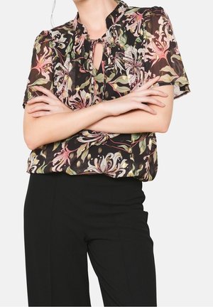 Femme portant un chemisier noir à manches courtes à motif floral avec un col à nouer, associé à un pantalon noir taille haute, bras croisés sur la poitrine.