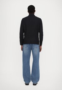 Schwarzes Hemd mit langen Ärmeln und Kragen, versehen mit Knopf-Details. Kombiniert mit blauen, weiten Jeans für eine entspannte Silhouette.