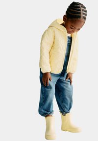 Ein Kind mit geflochtenen Haaren schaut nach unten und trägt eine gelbe Steppjacke, blaue Jeanslatzhose und helle gelbe Gummistiefel vor einem weißen Hintergrund.