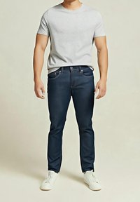 Homme portant une chemise à manches courtes gris clair, un jean bleu foncé et des baskets blanches, debout devant un fond uni.