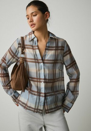 Frau trägt ein braun-blau kariertes Hemd mit Knöpfen und hellgraue Hosen, sie hat eine braune Wildleder-Umhängetasche dabei.