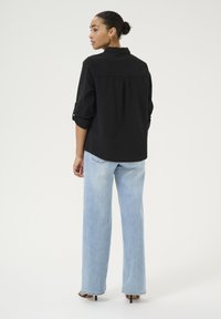 Femme avec un chignon portant une chemise à manches longues noire et un jean large bleu clair, debout et regardant à sa gauche.
