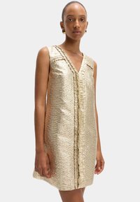 Vestido dorado sin mangas con una tela texturizada y arrugada. Presenta un escote en V y un detalle frontal con volantes, con un diseño minimalista.