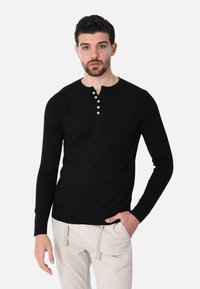 Maglione lungo nero stile henley con texture a costine e placca con cinque bottoni, indossato con pantaloni chiari.