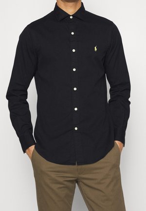 Mand iført en sort langærmet skjorte med knapper og en lille gul logo på brystet, samt brune bukser, hænderne i lommerne.