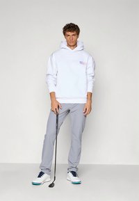 Sweatshirt branco com capuz, bolso frontal e logótipo, combinado com calças cinzentas. Modelo segura um taco de golfe, calçando sapatos brancos com detalhes azuis.