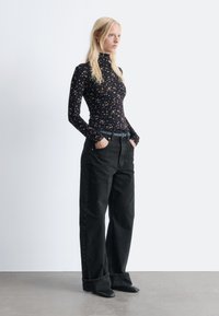 Long-sleeve black top with a floral pattern, paired with loose-fitting black jeans and pointed black shoes, standing against a neutral background.