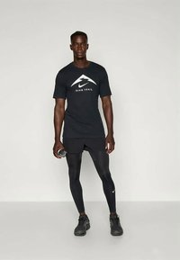 Must Nike Trail t-särk valge logoga, mustade kompressioonpükstega ja mustade tossudega; hoiab veepudelit.