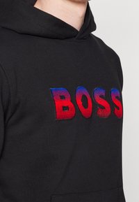 Felpa nera con cappuccio e logo "BOSS" in rilievo ricamato a sfumature rosse e blu. Presenta una tasca a marsupio e tessuto morbido.