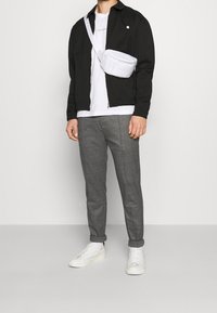 Svart lättjacka över en vit t-shirt, grå mönstrade byxor och vita sneakers. En vit axelväska bärs över jackan.