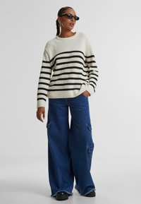 Maglione a righe bianche e nere con texture a costine, abbinato a pantaloni cargo in denim blu a gamba larga con tasche laterali e orli sfrangiati.